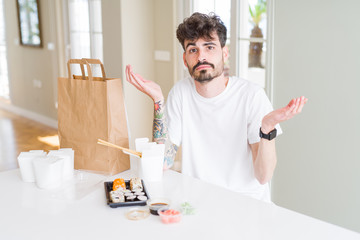 Young man eating asian sushi from home delivery clueless and confused expression with arms and hands raised. Doubt concept.