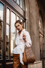 Outdoor fashion portrait of elegant, luxury woman wearing trendy white shirt, leather trousers, holding beige, brown reptile, snakeskin, python print bag, handbag, walking in street of European city