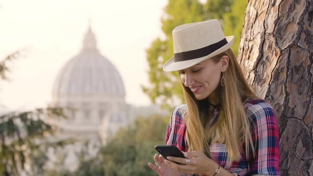 Happy young blonde woman with long hair and blue eyes laining on a tree, using her smartphone in front of Rome cityscape. Saint Peter church from Gianicolo panoramic view.