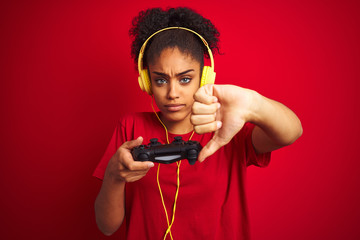 Afro woman playing video game using joystick and headphones over isolated red background with angry face, negative sign showing dislike with thumbs down, rejection concept