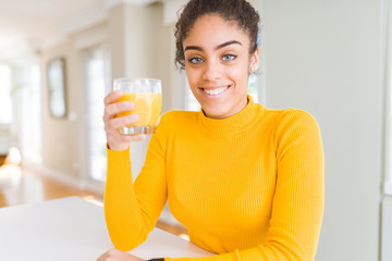 Young african american woman drinking a glass of fresh orange juice with a happy face standing and smiling with a confident smile showing teeth