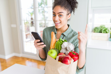 Young african american girl holding paper bag of groceries and using smartphone surprised with an...