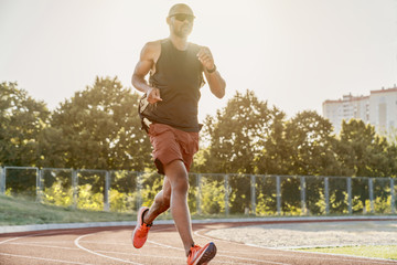 Young male athlete in sunglasses and earphones training on a race track. Afro sprinter running on...