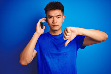 Young asian chinese man talking on the smartphone standing over isolated blue background with angry...