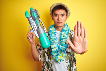 Asian chinese man wearing floral hawaian lei and water gun over isolated yellow background with...