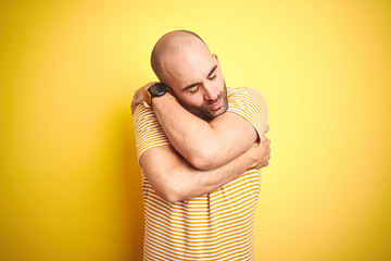 Young bald man with beard wearing casual striped t-shirt over yellow isolated background Hugging...