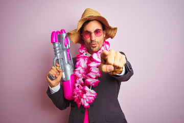 Young businessman wearing water gun hawaiian lei hat glasses over isolated pink background pointing...