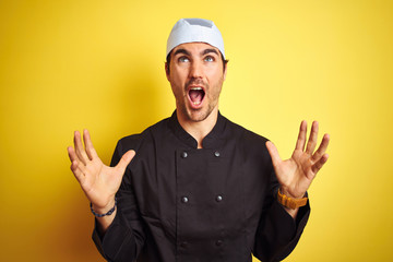 Young handsome chef man cooking wearing uniform and hat over isolated yellow background crazy and mad shouting and yelling with aggressive expression and arms raised. Frustration concept.