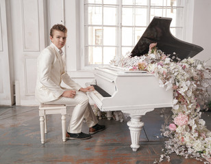 young man teenager in a white business suit with a large white piano in flowers. caucasian male play music at holiday alone.