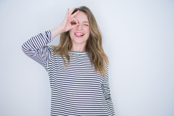 Beautiful blonde girl with blue eyes wearing striped sweater over white isolated background doing ok gesture with hand smiling, eye looking through fingers with happy face.