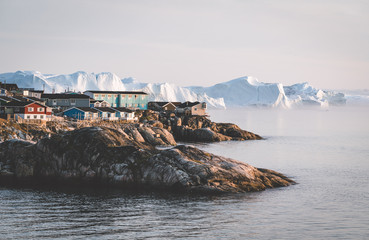 Aerial View of Arctic city of Ilulissat, Greenland during sunrise sunset with fog. Colorful houses in the center of the town with icebergs in the background in summer on a sunny day with orange pink © Mathias