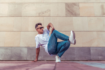 Young stylish athlete background wall, guy dancer, summer in city, dancing break dance, healthy fitness athlete life style, white t-shirt jeans sneakers. Dancing to the music. Performs public.
