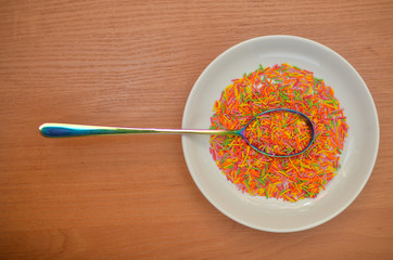 Long baking sprinkle in a white saucer with a rainbow spoon on a wooden table, top view