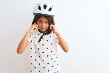 Beautiful child girl wearing security bike helmet standing over isolated white background covering ears with fingers with annoyed expression for the noise of loud music. Deaf concept.
