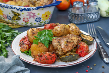 Chicken thighs baked with potatoes, tomatoes and zucchini, located in a plate and in ceramic form, on a dark background, horizontal orientation