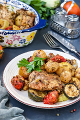 Chicken thighs baked with potatoes, tomatoes and zucchini, located in a plate and in ceramic form, on a dark background, vertical orientation