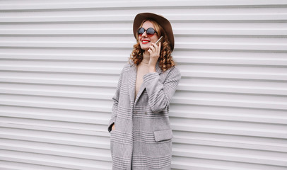 Portrait elegant smiling woman calling on smartphone wearing gray coat, round hat looking away over white wall background