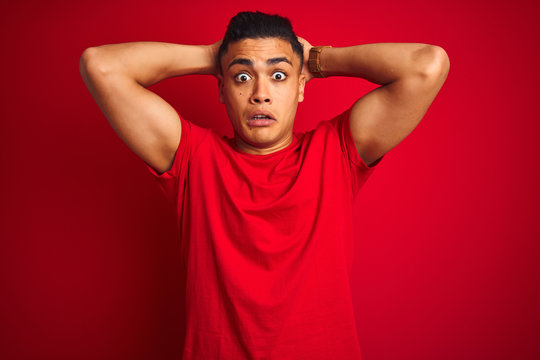 Young brazilian man wearing t-shirt standing over isolated red background Crazy and scared with hands on head, afraid and surprised of shock with open mouth