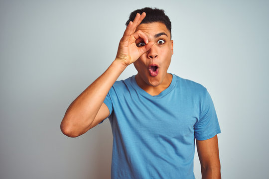 Young brazilian man wearing blue t-shirt standing over isolated white background doing ok gesture shocked with surprised face, eye looking through fingers. Unbelieving expression.