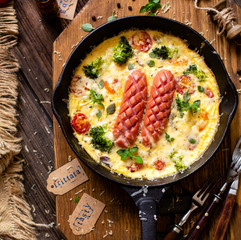 overhead shot of homemade tasty baked omelette or frittata with two sausages, broccoli, cherry tomatoes, basil, melted cheese in black skillet on wooden board on rustic table with sackcloth