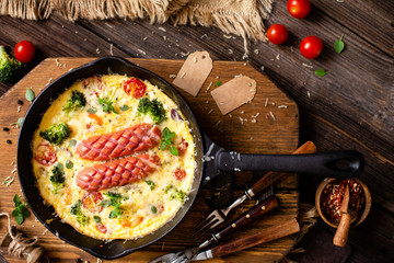 overhead shot of homemade tasty baked omelette or frittata with two sausages, broccoli, cherry tomatoes, basil, melted cheese in black skillet on wooden board on rustic table with sackcloth