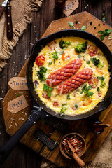 overhead shot of homemade tasty baked omelette or frittata with two sausages, broccoli, cherry tomatoes, basil, melted cheese in black skillet on wooden board on rustic table with sackcloth