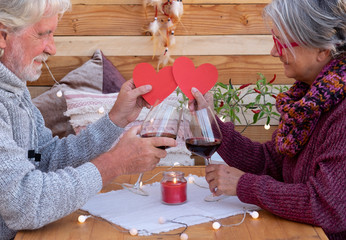 A smiling senior couple show two red heart shapes. sitting outdoor in a romantic corner made by wood. Yellow lights. Two red wineglasses to celebrate a positive moment