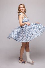 Naklejka premium Full length portrait of positive beautiful lady in summer dress and sandals dancing with her skirt flying with plaster on her broken leg. Isolate on grey. Smiling woman dancing with plaster.