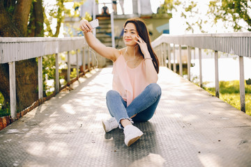 beautiful and bright girl in pink t-shirts and blue jeans sitting in the sunny summer park and use the phone with headphones