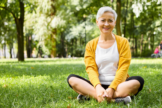 Positive friendly looking Caucasian female pensioner sitting on grass in stylish sportswear and sneakers, keeping her legs crossed, having rest after cardio training on warm September day, smiling