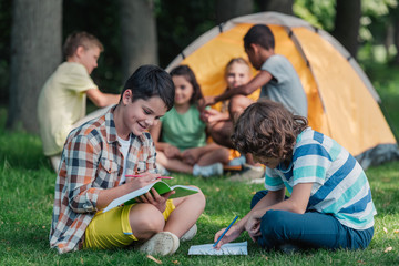 selective focus of happy boys writing on notebooks near multicultural friends in camp