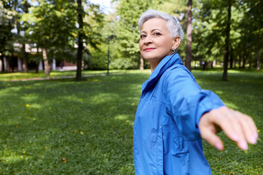Follow me. Outdoor image of happy beautiful gray haired mature female posing in wild nature turning back with outstretched arm, making inviting gesture, going to guide though forest. hand out of focus