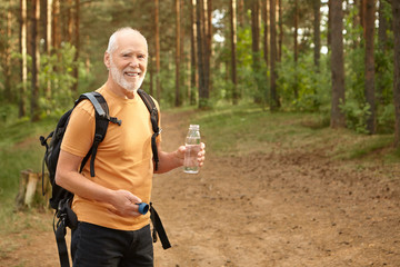 Cheerful attractive senior man with backpack hiking outdoors, smiling joyfully satisfying his thirst, holding bottle with drinking water, posing in pine forest. Age, maturity and active lifestyle