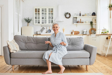 Indoor shot of joyful charming elderly housewife in stylish dress sitting on large gray couch with...
