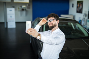 Visiting car dealership. Handsome man is doing selfie with his new car