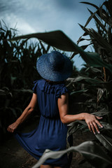 Beautiful woman with blue hat and dress from behind posing in corn field during stormy clouds.