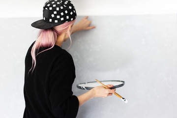 Back view of young girl with pink hair, painting wall with brush and white color, in apartment.