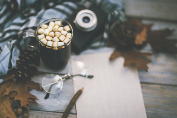 Autumn background on a wooden table with a knitted wool shawl, an old camera, autumn oak leaves and acorns, coffee in a glass mug and a notepad with a pencil. Cozy warm mockup