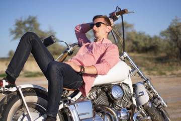 Naklejka premium Young stylish motorcyclist driving his motorbike in the desert road during sunset