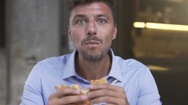 Funny close up of hungry starving handsome and young businessman eating devouring a big hamburger during lunch break. Eating chips and drinking a beer.