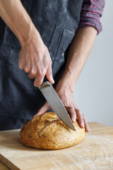 Male baker cuts sourdough bread on a wooden table, close-up, isolated on a light background. Natural light from the window