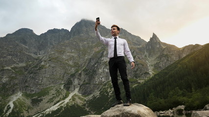 Successful confident businessman talking by video chat, uses a mobile phone on online conference near a lake in the rocky mountains at sunset, way to success