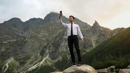 Obraz premium Successful confident businessman talking by video chat, uses a mobile phone on online conference near a lake in the rocky mountains at sunset, way to success