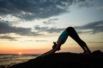 A young woman practicing yoga during sunset on the seashore. Asana downward dog.