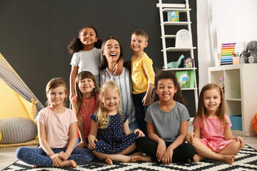 Cute little children with nursery teacher sitting on floor in kindergarten. Indoor activity