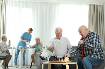 Elderly men playing chess while nurse serving breakfast to women at retirement home. Assisting senior people