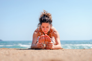 Young woman in summer dress practice yoga at the beach by the sea or ocean in sunny day stretching paschimottanasana