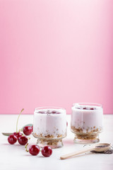 Yoghurt with cherries, chia seeds and granola in glass with wooden spoon on pink and white background. side  view