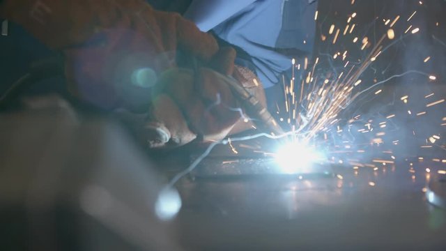 Close-up blacksmith welder in protective mask works with metal using a welding machine, bright sparks and flashes in super slow motion