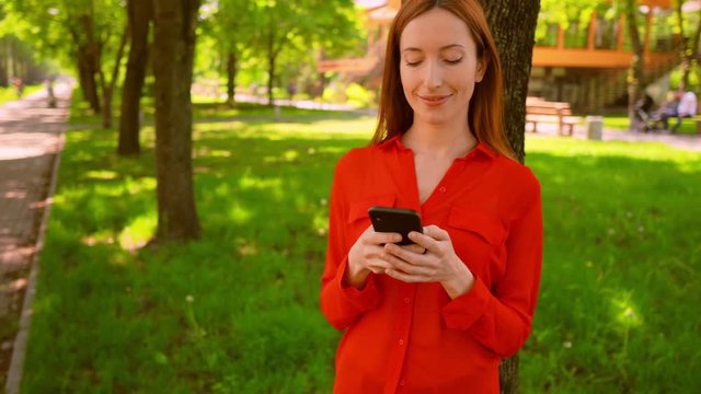 adult woman with red hair holding cellphone typing message or chatting online standing in the park near tree summer outdoors background
